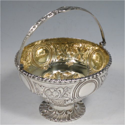 An Antique Victorian Sterling Silver sweet-meat basket, having a round baluster body with gold-gilt interior and an applied gadroon border, with hand-chased floral decoration between four round vacant cartouches, a hinged and florally chased swing handle, and all sitting on a swirl-fluted pedestal foot. Made by Martin and Hall of London in 1871. The dimensions of this fine hand-made antique silver basket are height 15 cms (6 inches), diameter 12.5 cms (5 inches), and it weighs approx. 224g (7.2 troy ounces).