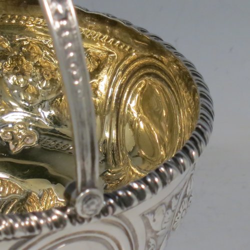 An Antique Victorian Sterling Silver sweet-meat basket, having a round baluster body with gold-gilt interior and an applied gadroon border, with hand-chased floral decoration between four round vacant cartouches, a hinged and florally chased swing handle, and all sitting on a swirl-fluted pedestal foot. Made by Martin and Hall of London in 1871. The dimensions of this fine hand-made antique silver basket are height 15 cms (6 inches), diameter 12.5 cms (5 inches), and it weighs approx. 224g (7.2 troy ounces).