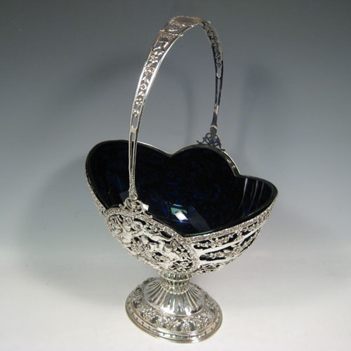 Antique Victorian German silver flower basket, having a hand-pierced body with hand-chased floral and cherub decoration, a lobed blue-glass liner, swing handle, and sitting on a pedestal foot. Made in Germany in ca. 1880. Height inc. handle 41 cms (16 inches), length 24 cms (9.5 inches), width 18 cms (7 inches). Weight approx. 765g (24.7 troy ounces). Please note that this piece is made from 800 standard silver.