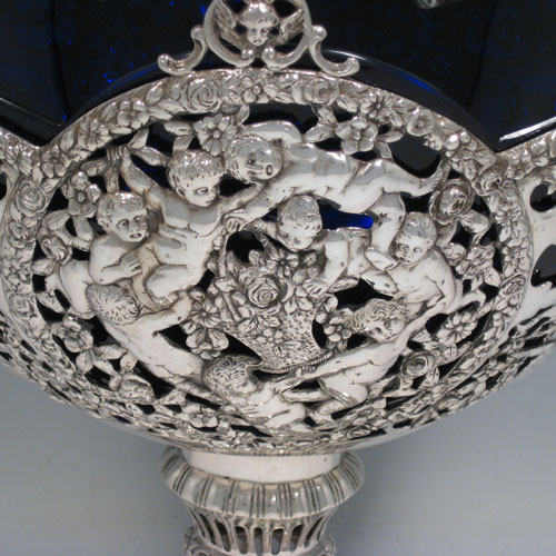 Antique Victorian German silver flower basket, having a hand-pierced body with hand-chased floral and cherub decoration, a lobed blue-glass liner, swing handle, and sitting on a pedestal foot. Made in Germany in ca. 1880. Height inc. handle 41 cms (16 inches), length 24 cms (9.5 inches), width 18 cms (7 inches). Weight approx. 765g (24.7 troy ounces). Please note that this piece is made from 800 standard silver.