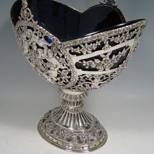 Antique Victorian German silver flower basket, having a hand-pierced body with hand-chased floral and cherub decoration, a lobed blue-glass liner, swing handle, and sitting on a pedestal foot. Made in Germany in ca. 1880. Height inc. handle 41 cms (16 inches), length 24 cms (9.5 inches), width 18 cms (7 inches). Weight approx. 765g (24.7 troy ounces). Please note that this piece is made from 800 standard silver.