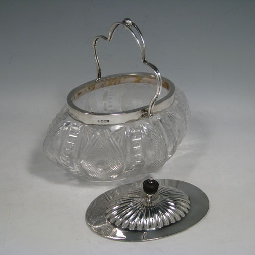 A very pretty Antique Victorian Sterling Silver and hand-cut crystal biscuit jar, having an unusual oval crystal body with hand-cut floral and geometric work, a lift-off lid with half-fluting and wooden finial, and a wire-work swing handle. Made by Fordham and Faulkner of Sheffield in 1901. The dimensions of this fine hand-made antique silver and crystal biscuit jar are height 18 cms (7 inches), length 20 cms (8 inches), and width 15 cms (6 inches).