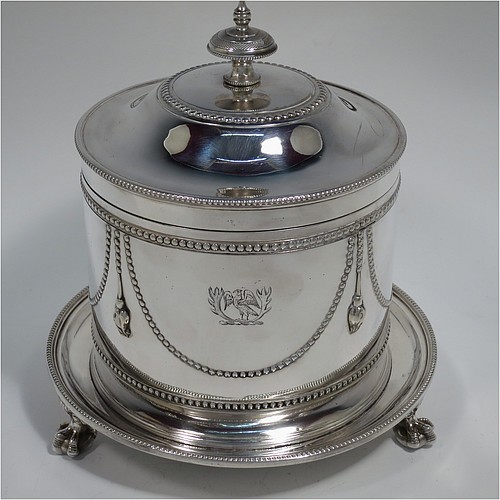 An Antique Victorian Silver Plated biscuit box, having a round body with applied bead-edged borders, straight sides and hand-chased swags of bead-work in a neoclassical style, a hinged domed lid with cast finial, and all sitting on an attached plate with three cast claw and ball feet. Made by James Thompson of Sheffield in ca. 1880. The dimensions of this fine hand-made antique silver-plated biscuit box are height 18 cms (7 inches), and diameter at base 18 cms (7 inches). Please note that this item is crested.