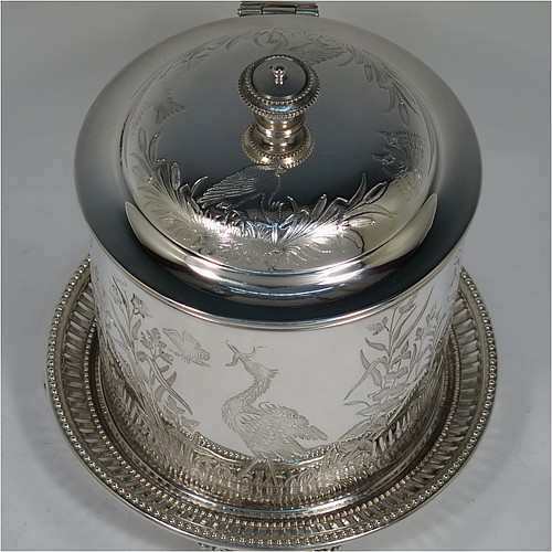 An Antique Victorian Silver Plated Chinoiserie style biscuit box, having a round body with straight sides and applied bead-edged borders, with very pretty hand-engraved flowers, reeds, birds, and butterfly work, a hinged domed lid with cast finial, and all sitting on a hand-pierced attached plate with four cast scroll feet. Made by the Atkin Brothers of Sheffield in ca. 1880. The dimensions of this fine hand-made antique silver-plated biscuit box are height 18 cms (7 inches), and diameter at base 17.5 cms (7 inches).    