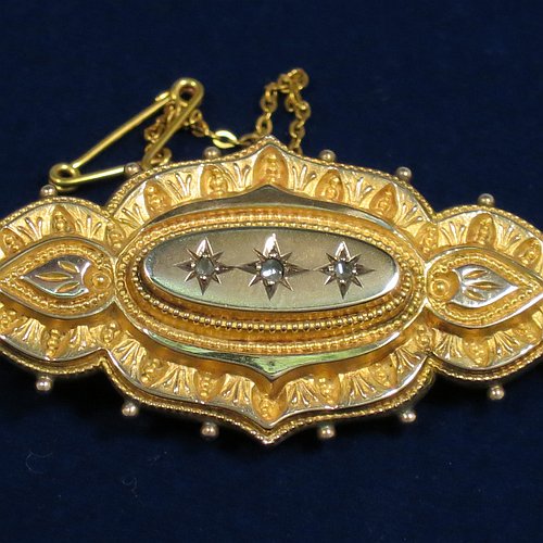 An Antique Victorian Etruscan style 9ct carat gold brooch, having an elongated and shaped oval body, with applied bead decoration, and a stepped oval centre with three old rose-cut diamonds, and the back having a pin with oval locket and glass cover, and an attached security chain. Made in ca. 1880. The dimensions of this fine antique gold brooch are length 5 cms (2 inches), width 2.5 cms (1 inch), and it weighs approx. 7g.