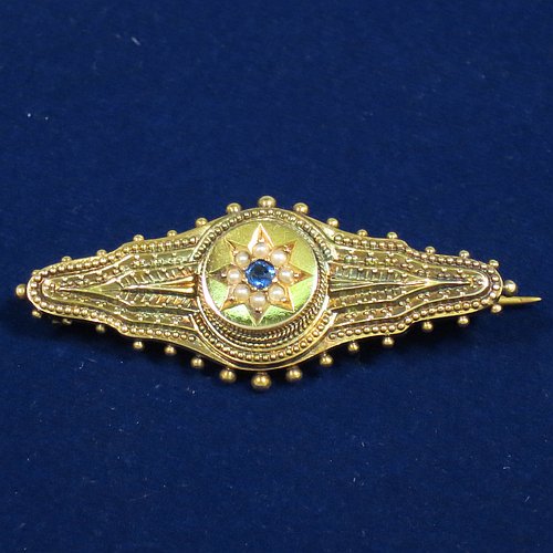 An Antique Victorian Etruscan style 15ct carat gold brooch, having an elongated and shaped oval body, with applied bead decoration, and a stepped round centre with star decoration having a sapphire and small pearls, and the back having a pin and a security ring but no chain. Made in ca. 1880. The dimensions of this fine antique gold brooch are length 4.5 cms (1.75 inches), width 2 cms (0.75 inch), and it weighs approx. 3.5g.   