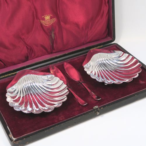 An elegant Antique Victorian Sterling Silver pair of butter shells, having traditional scallop shapes, and sitting on three cast ball feet, together with a pair of original butter knives in the Old English pattern, and all sitting in an original maroon satin and velvet-lined presentation box. This handsome pair of silver butter shells and knives were all made by the Atkin Brothers of Sheffield in 1900. The dimensions of this fine hand-made set of antique silver butter shells and knives are length 12 cms (4.75 inches), width 10 cms (4 inches), with a total weight of approx. 136g (4.4 troy ounces).  