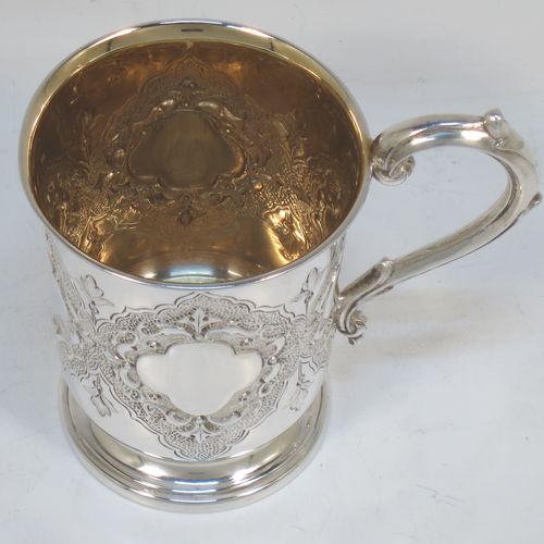 A very pretty Antique Victorian Sterling Silver christening mug, having a round body with straight tapering sides and tucked under belly, with hand-chased floral and scroll-work decoration surrounding four vacant lobed cartouches, a scroll loop handle, a gold-gilt interior, and all sitting on a pedestal foot. Made by Robert Hennell of London in 1865. The dimensions of this fine hand-made antique silver christening mug are height 10 cms (4 inches), diameter at lip 7 cms (2.75 inches), and it weighs approx. 130g (4.2 troy ounces).  