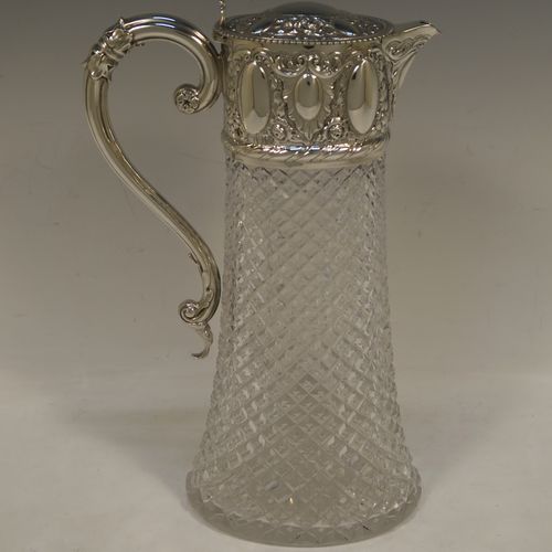 A very pretty Antique Victorian Sterling Silver and hand-cut crystal claret jug, having a hand-chased mount with floral and scroll decoration, with three oval vacant cartouches on both sides, a hinged domed lid with a gadroon border and finial, a scrolled handle with leaf work, and an oval hand-cut straight-sided body with hobnail pattern and star-cut base. Made by William Hutton of London in 1895. The dimensions of this fine hand-made antique silver and crystal claret jug are height 27 cms (10.75 inches), and length 17 cms (6.75 inches).  