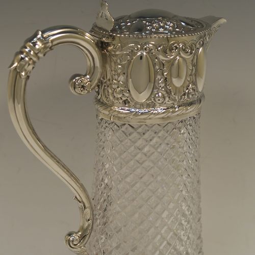 A very pretty Antique Victorian Sterling Silver and hand-cut crystal claret jug, having a hand-chased mount with floral and scroll decoration, with three oval vacant cartouches on both sides, a hinged domed lid with a gadroon border and finial, a scrolled handle with leaf work, and an oval hand-cut straight-sided body with hobnail pattern and star-cut base. Made by William Hutton of London in 1895. The dimensions of this fine hand-made antique silver and crystal claret jug are height 27 cms (10.75 inches), and length 17 cms (6.75 inches).  