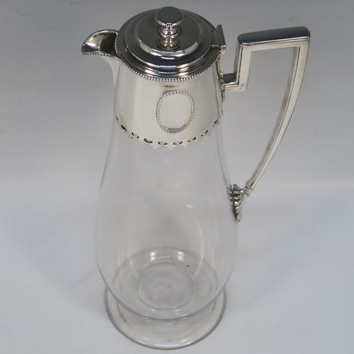 A very elegant Antique Victorian Sterling Silver and hand-cut crystal claret jug, having a plain round mount with applied bead-edged borders, a plain square cross-section side handle, a hinged stepped lid with a cast round finial, together with a hand-cut crystal round body with straight tapering sides and a tucked under belly above a pedestal foot. This handsome silver and crystal claret jug was made by Charles Favell of Sheffield in 1875. The dimensions of this fine hand-made antique silver and crystal claret jug are height 28 cms (11 inches), and length 13.5 cms (5.25 inches). Please note that this item has two oval cartouches with bead borders one of which is  engraved with a part crest.   