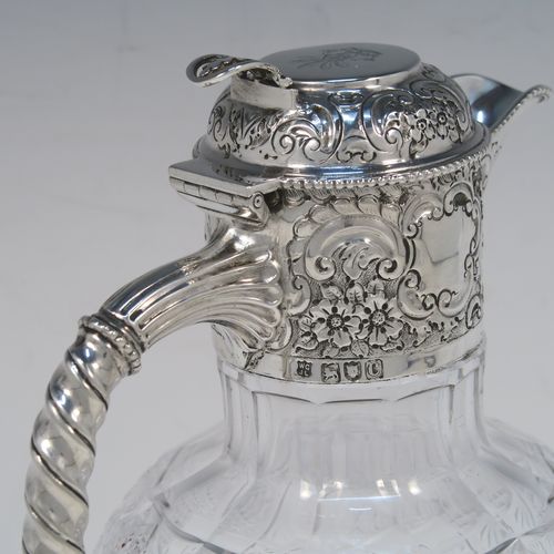 A very beautiful Antique Victorian Sterling Silver and hand-cut crystal claret jug, having an ornate round mount with hand-chased floral and scroll decoration, a fluted and twisted style side handle, a hinged lid with matching decoration and a pierced thumb-piece, together with a hand-cut crystal round body with tapering sides and above a flat star-cut base. This beautiful antique silver and crystal claret jug was made by Gibson and Langland of London in 1898. The dimensions of this fine hand-made antique silver and crystal claret jug are height 23 cms (9 inches), and length 14 cms (5.5 inches). Please note that this item is crested on the top of the lid.