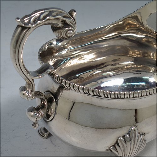 An Antique Georgian Sterling Silver pair of large and heavy George III sauce or gravy boats, having plain bellied bodies, with applied gadroon borders, cast scroll handles with anthemion leaf thumb-pieces, and sitting on three cast hoof feet with shell shoulders. Made by Thomas Wallis I of London in 1773. The dimensions of these fine hand-made antique silver gravy or sauce boats are length 18.5 cms (7.25 inches), width 10 cms (4 inches), height 14 cms (5.5 inches), and they weigh a total of 821g (26.5 troy ounces). Please note that these items are crested.