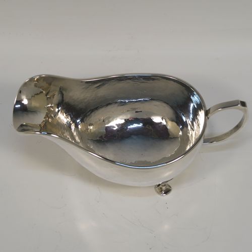 A Sterling Silver cream or sauce boat in an Arts and Crafts style, having a bellied body with hand-hammered or Planished decoration, a scroll side-handle, and sitting on three cast and panelled feet. This elegant silver sauce or cream boat was made by A. E. Jones of Birmingham in 1928. The dimensions of this fine hand-made silver cream or sauce boat are length 17 cms (6.75 inches), height 8 cms (3 inches), width 8.5 cms (3.25 inches), and it weighs approx. 162g (5.2 troy ounces).   