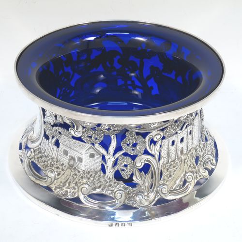 A very beautiful Irish Antique Edwardian Sterling Silver large table dish ring, having a hand-pierced and chased body decorated with scrolls, two cottages, a castle, and a bridge, a vacant oval cartouche, and a blue-glass liner. Made by Edmund Johnson of Dublin in 1910. The dimensions of this fine hand-made antique silver dish ring are height 9 cms (3.5 inches), diameter at base 19 cms (7.5 inches), and it weighs approx. 302g (9.7 troy ounces).  