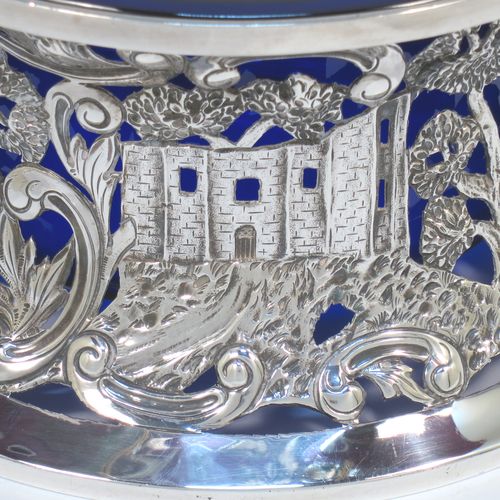 A very beautiful Irish Antique Edwardian Sterling Silver large table dish ring, having a hand-pierced and chased body decorated with scrolls, two cottages, a castle, and a bridge, a vacant oval cartouche, and a blue-glass liner. Made by Edmund Johnson of Dublin in 1910. The dimensions of this fine hand-made antique silver dish ring are height 9 cms (3.5 inches), diameter at base 19 cms (7.5 inches), and it weighs approx. 302g (9.7 troy ounces).  