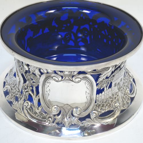 A very beautiful Irish Antique Edwardian Sterling Silver large table dish ring, having a hand-pierced and chased body decorated with scrolls, two cottages, a castle, and a bridge, a vacant oval cartouche, and a blue-glass liner. Made by Edmund Johnson of Dublin in 1910. The dimensions of this fine hand-made antique silver dish ring are height 9 cms (3.5 inches), diameter at base 19 cms (7.5 inches), and it weighs approx. 302g (9.7 troy ounces).  