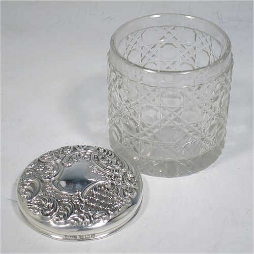 An Antique Victorian Sterling Silver and cut-crystal dressing table jar / box, having a round hand-cut crystal body in the Hobnail pattern, a hand-chased pull-off lid with floral decoration, and sitting on a flat star-cut base. Made by Adie & Lovekin Ltd., of Birmingham in 1901. The dimensions of this fine hand-made antique silver and crystal dressing table box / jar are height 8 cms (3 inches), and diameter 7 cms (2.75 inches).   