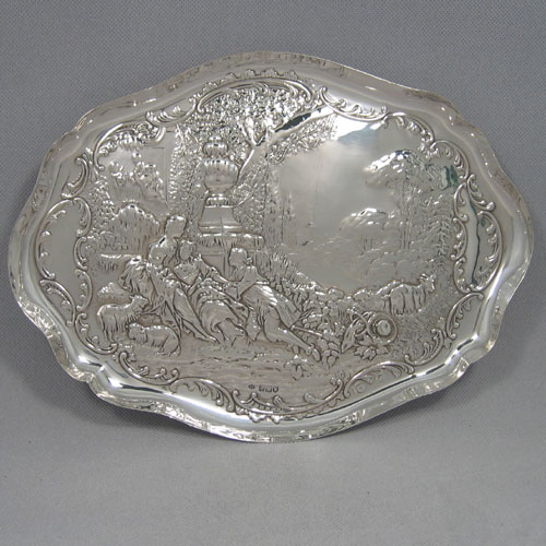 An Antique Edwardian Sterling Silver dressing table pin tray, having a shaped oval body with a raised edge border, and a hand-chased pastoral scene of figures reclining under a spreading oak tree, and all sitting on a flat base. Made by William Comyns of London in 1907. The dimensions of this fine hand-made antique silver pin or dressing table tray are length 31 cms (12 inches), width 23 cms (9 inches), and it weighs approx. 341g (11 troy ounces).