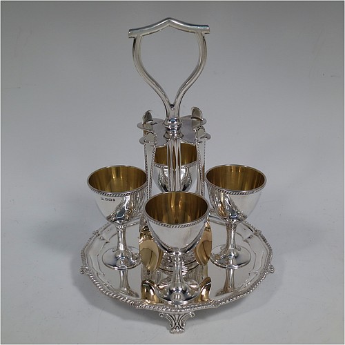 An Antique Victorian Sterling Silver egg cruet set, having four plain round egg cups with gold-gild interiors sitting on pedestal feet, four egg spoons with gold-gilt bowls, and a plain cast loop handle attached to a round base with an applied shaped gadroon border, and sitting on four cast scroll feet. Made by George Hancock of Sheffield in 1897 (the spoons by James Dixon and Sons of Sheffield in 1917). The dimensions of this fine hand-made antique silver egg cruet set are height 20 cms (8 inches), diameter of base is 15 cms (6 inches), and the total weight is approx. 688g (22 troy ounces).   