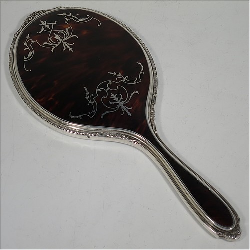 A Sterling Silver and Tortoise-shell hand-mirror, having an oval body with an applied floral border, with very pretty Neoclassical style inlaid tortoiseshell decoration, and an original bevelled mirror. Made by Mappin and Webb of Birmingham in 1928. The dimensions of this fine hand-made silver and tortoise-shell hand mirror are length 28 cms (11 inches), and width of mirror 11 cms (4.25 inches).    