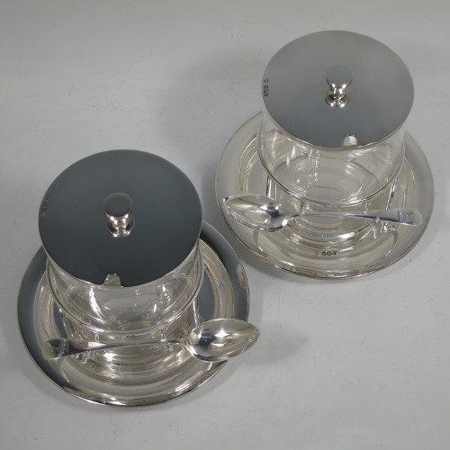 A very unusual, large, and handsome pair of Antique Victorian sterling silver and hand-cut crystal jam, honey, or preserve jars, having round straight-sided bodies with plain pull-off lids with disc finials, and sitting in gallery frames with two hooks for holding jam or honey spoons, and attached to round base plates with three cushion feet underneath. Made by Charles Edward Nixon of Sheffield in 1896. The dimensions of these fine hand-made silver and crystal jam, honey, or conserve jars are height 13.5 cms (5.25 inches), diameter at base 15 cms (6 inches) square. Please note that these come with two matching silver serving spoons.  