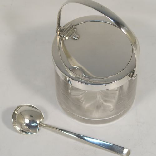 A very handsome Antique Edwardian Sterling Silver and crystal honey or jam jar, having a plain cylindrical body with star-cut base, a plain round mount, with a mechanical swing-handled hinged lid. Together with a matching plain honey spoon. This beautiful honey or jam jar was made by Goldsmiths & Silversmiths of London in 1909, and the spoon was made by John Grinsell and Sons of Birmingham in 1910. The dimensions of this fine hand-made antique silver and glass conserve jar are height (inc. handle) 12 cms (4.75 inches), and diameter 8 cms (3 inches).  