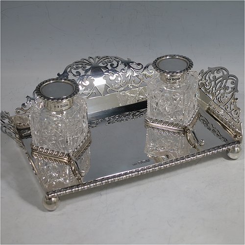 A very pretty Antique Edwardian Sterling Silver galleried double inkstand, having a rectangular body with three hand-pierced scroll-work gallery sides, an applied gadroon front border, two square hand-cut crystal removable ink bottles with round hinged lids, together with two applied pen-rest scrolls, all sitting on four ball feet. Made by George Wish of Sheffield in 1905. The dimensions of this fine hand-made antique silver double inkstand are length 22 cms (8.75 inches), width 14 cms (5.5 inches), height 5 cms (2 inches), and it weighs approx. 334g (10.8 troy ounces).   