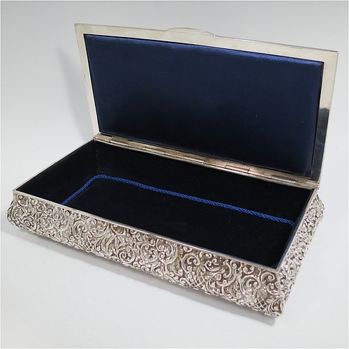 An Antique Edwardian Sterling Silver large casket style jewellery dressing table box, having a rectangular shaped casket style body, with hand-chased floral decoration, a hinged lid with a central vacant cartouche and a plain border with thumb-piece, a dark blue velvet and satin-lined interior, all sitting on a flat base. Made by Henry Matthews of Birmingham in 1902. The dimensions of this fine hand-made antique silver dressing table jewellery box are length 30 cms (11.75 inches), width 16 cms (6.25 inches), and height 6.5 cms (2.5 inches).   