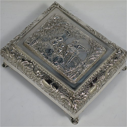 A very pretty Antique Victorian Sterling Silver casket style jewellery dressing table box, having a rectangular shaped casket style body, with hand-chased floral and scroll decoration, a hinged lid with a hand-chased figural scene, a dark green velvet-lined interior, and all sitting on four shell feet. Made by Henry Matthews of Birmingham in 1900. The dimensions of this fine hand-made antique silver dressing table jewellery box are length 16 cms (6.25 inches), width 13.5 cms (5.25 inches), and height 7 cms (2.75 inches).    