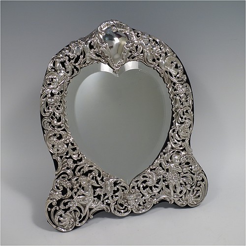 An Antique Victorian Sterling Silver table or vanity mirror having hand-chased cherubs and birds, scroll-work, and floral decoration, with a top vacant cartouche, an original bevelled heart-shaped mirror, and blue velvet backing with easel stand. Made by William Comyns of London in 1893. The dimensions across the frame of this fine hand-made antique silver vanity or table mirror are height 28 cms (11 inches), and width 24 cms (9.5 inches).    