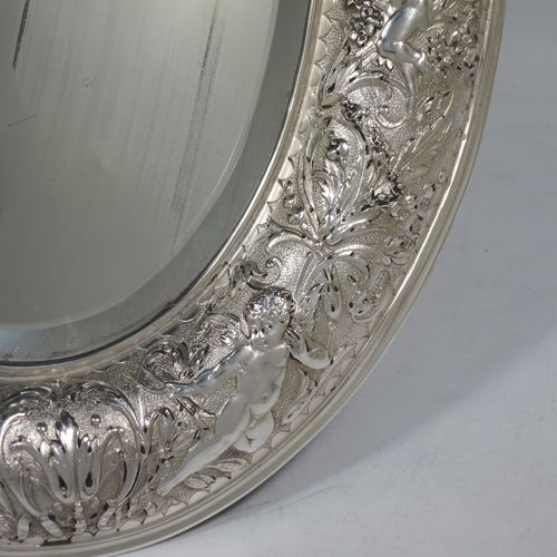 A very unusual and pretty Antique Victorian Sterling Silver table or vanity mirror (that also doubles as a mirror plateau), having hand-chased cherubs and floral work, surrounding an original bevelled oval mirror, and a blue velvet backing with eight stud feet and an easel stand. Made by Elkington and Co., of Birmingham in 1900. The dimensions across the frame of this fine hand-made antique silver vanity or table mirror are height 38 cms (15 inches), and width 27 cms (10.5 inches). Please note that the silver backing to the mirror has age-related marks, but nothing too serious.   