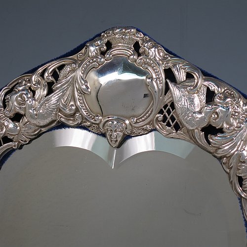 An Antique Victorian Sterling Silver table or vanity mirror having hand-chased birds, scroll-work, and floral decoration, with a top vacant cartouche, an original bevelled heart-shaped mirror, and blue velvet backing with easel stand. Made by William Comyns of London in 1897. The dimensions across the frame of this fine hand-made antique silver vanity or table mirror are height 28 cms (11 inches), and width 24 cms (9.5 inches).   