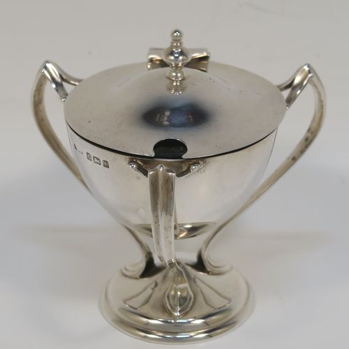 A very stylish Antique Edwardian Art Nouveau mustard pot, having a plain round bellied body, with a hinged lid and a cast urn finial, and three scroll side-handles attached to a round pedestal foot, and with an internal blue glass liner. This silver Art Nouveau mustard pot was made by Edward Souter Barnsley of Birmingham in 1905. The dimensions of this fine hand-made antique silver mustard pot are height 8 cms (3 inches), diameter 5.5 cms (2.25 inches), and it weighs approx. 83g (2.7 troy ounces).   