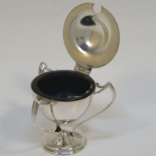 A very stylish Antique Edwardian Art Nouveau mustard pot, having a plain round bellied body, with a hinged lid and a cast urn finial, and three scroll side-handles attached to a round pedestal foot, and with an internal blue glass liner. This silver Art Nouveau mustard pot was made by Edward Souter Barnsley of Birmingham in 1905. The dimensions of this fine hand-made antique silver mustard pot are height 8 cms (3 inches), diameter 5.5 cms (2.25 inches), and it weighs approx. 83g (2.7 troy ounces).   