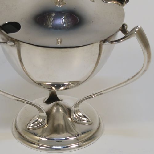 A very stylish Antique Edwardian Art Nouveau mustard pot, having a plain round bellied body, with a hinged lid and a cast urn finial, and three scroll side-handles attached to a round pedestal foot, and with an internal blue glass liner. This silver Art Nouveau mustard pot was made by Edward Souter Barnsley of Birmingham in 1905. The dimensions of this fine hand-made antique silver mustard pot are height 8 cms (3 inches), diameter 5.5 cms (2.25 inches), and it weighs approx. 83g (2.7 troy ounces).   