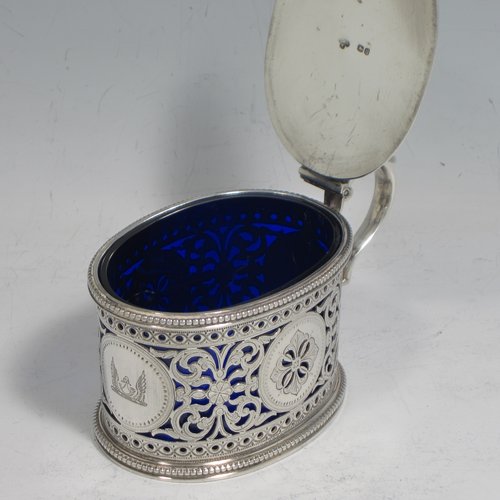 Antique Victorian sterling silver mustard pot, having an oval straight-sided body with beautiful hand-pierced and engraved floral decoration, with applied bead borders, a hinged flat lid with hand-pierced thumb-piece, a scroll handle and a blue glass liner. Made by the Barnard Brothers of London in 1875. The dimensions of this fine hand-made silver mustard pot are height 7 cms (2.75 inches), length 11 cms (4.25 inches), and it weighs approx. 140g (4.5 troy ounces). Please note that this item is crested and monogrammed.   