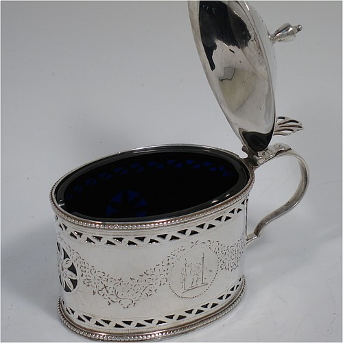 
An Antique Georgian Neoclassical Sterling Silver mustard pot, having an oval body with straight sides, with hand-pierced and engraved neoclassical style floral decoration, a hinged and domed lid with urn shaped finial and pierced thumb-pierce, a scroll handle, a blue glass liner, and all sitting on a flat base. Made by Robert Hennell I  of London in 1773. The dimensions of this fine hand-made antique silver mustard pot are height 8 cms (3.25 inches), length 10 cms (4 inches), width 5.5 cms (2.25 inches), and it weighs approx. 93g (3 troy ounces).    