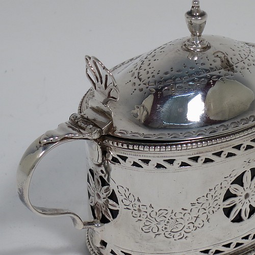 
An Antique Georgian Neoclassical Sterling Silver mustard pot, having an oval body with straight sides, with hand-pierced and engraved neoclassical style floral decoration, a hinged and domed lid with urn shaped finial and pierced thumb-pierce, a scroll handle, a blue glass liner, and all sitting on a flat base. Made by Robert Hennell I  of London in 1773. The dimensions of this fine hand-made antique silver mustard pot are height 8 cms (3.25 inches), length 10 cms (4 inches), width 5.5 cms (2.25 inches), and it weighs approx. 93g (3 troy ounces).    