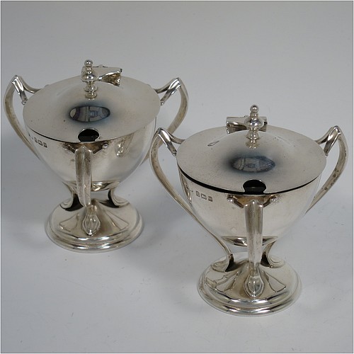 A very stylish Antique Edwardian Art Nouveau pair of mustard pots, having plain round bellied bodies, with hinged lids having cast urn finials, and three scroll side-handles attached to round pedestal feet, and with internal blue glass liners. Made by Edward Souter Barnsley of Birmingham in 1905. The dimensions of these fine hand-made antique silver mustard pots are height 8 cms (3 inches), diameter 5.5 cms (2.25 inches), and they weigh at total of approx. 164g (5.3 troy ounces).   