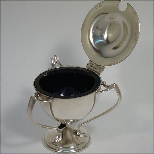 A very stylish Antique Edwardian Art Nouveau pair of mustard pots, having plain round bellied bodies, with hinged lids having cast urn finials, and three scroll side-handles attached to round pedestal feet, and with internal blue glass liners. Made by Edward Souter Barnsley of Birmingham in 1905. The dimensions of these fine hand-made antique silver mustard pots are height 8 cms (3 inches), diameter 5.5 cms (2.25 inches), and they weigh at total of approx. 164g (5.3 troy ounces).   