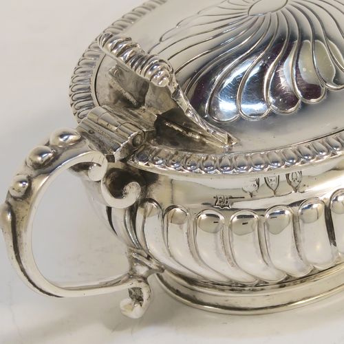 A beautiful Antique Victorian Sterling Silver mustard pot, having an oval bellied body with hand-chased fluted decoration, a gadroon-edged border, a hinged lid with cast scroll thumb-piece, a blue glass liner and scroll handle, all sitting on a collet foot. This very pretty antique silver mustard pot was made by Edward Hutton of London in 1891. The dimensions of this fine hand-made silver mustard pot are height 5.5 cms (2.25 inches), length 10.5 cms (4.25 inches), width 6.5 cms (2.5 inches), and it weighs approx. 142g (4.6 troy ounces).   