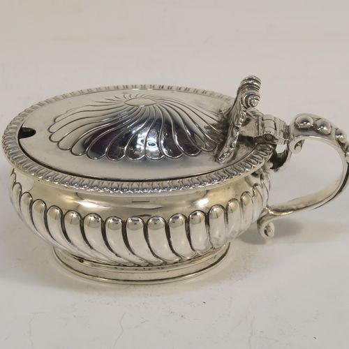 A beautiful Antique Victorian Sterling Silver mustard pot, having an oval bellied body with hand-chased fluted decoration, a gadroon-edged border, a hinged lid with cast scroll thumb-piece, a blue glass liner and scroll handle, all sitting on a collet foot. This very pretty antique silver mustard pot was made by Edward Hutton of London in 1891. The dimensions of this fine hand-made silver mustard pot are height 5.5 cms (2.25 inches), length 10.5 cms (4.25 inches), width 6.5 cms (2.5 inches), and it weighs approx. 142g (4.6 troy ounces).   