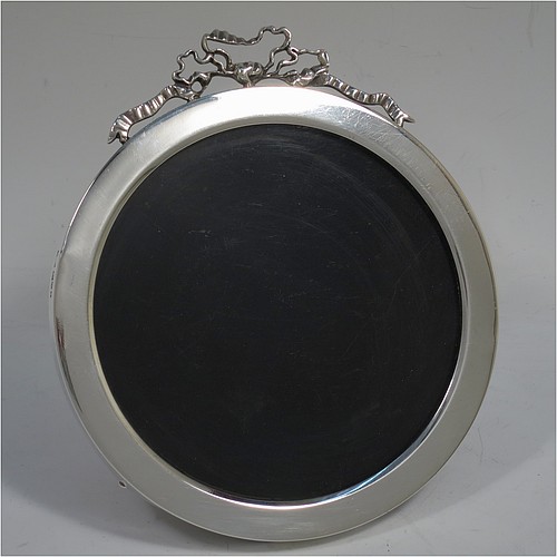 A very pretty Antique Edwardian Sterling Silver  photograph frame, having a plain round body with straight sides, an applied cast ribbon and bow top, and a brown wood-backed easel frame. Made by Stokes and Ireland Ltd., of Chester in 1905. The internal dimensions of this fine hand-made antique silver photo frame are diameter 11 cms (4.25 inches).    