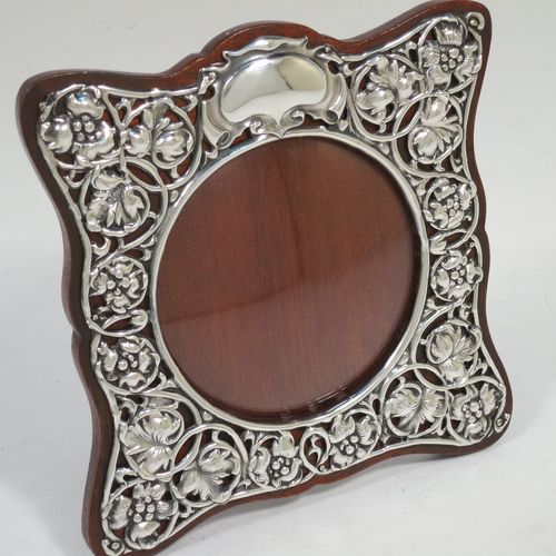 A very pretty Art Nouveau Antique Edwardian Sterling Silver photograph frame, having a round aperture with a convex glass cover, surrounded by a hand-chased and pierced border with floral and scroll decoration, and all pinned to a brown wood-backed easel frame. Made by Synyer and Beddoes of Birmingham in 1903. The internal dimensions of this fine hand-made Art Nouveau antique silver photo frame are 8.5 cms (3.3 inches) in diameter.   