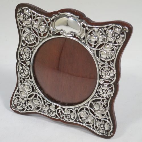 A very pretty Art Nouveau Antique Edwardian Sterling Silver photograph frame, having a round aperture with a convex glass cover, surrounded by a hand-chased and pierced border with floral and scroll decoration, and all pinned to a brown wood-backed easel frame. Made by Synyer and Beddoes of Birmingham in 1903. The internal dimensions of this fine hand-made Art Nouveau antique silver photo frame are 8.5 cms (3.3 inches) in diameter.   