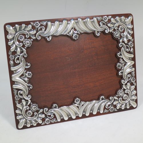 A very pretty Antique Victorian Sterling Silver photograph frame, having a rectangular landscape aperture, surrounded by a hand-pierced border with fluting, scroll-work, and floral decoration, and all pinned to a wood-backed easel frame. Made by William Comyns of London in 1895. The internal dimensions of this fine hand-made antique silver photo frame are 19 cms (7.5 inches) wide by 14 cms (5.5 inches) high.    