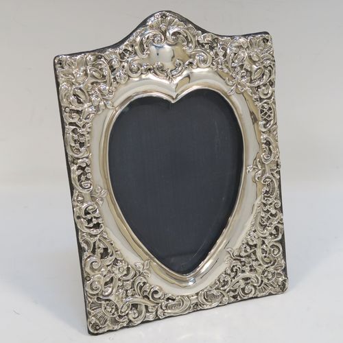 A very pretty Antique Edwardian Sterling Silver photograph frame, having a heart-shaped aperture, surrounded by a hand-chased body with floral and scroll decoration, and an original dark blue velvet-backed easel frame. This beautiful antique silver photo frame was made by James Deakin of Chester in 1902. The internal dimensions of this fine hand-made antique silver photo frame are 11.5 cms (4.5 inches) high by 9 cms (3.5 inches) wide. Please note that the velvet backing is slightly worn and faded.   