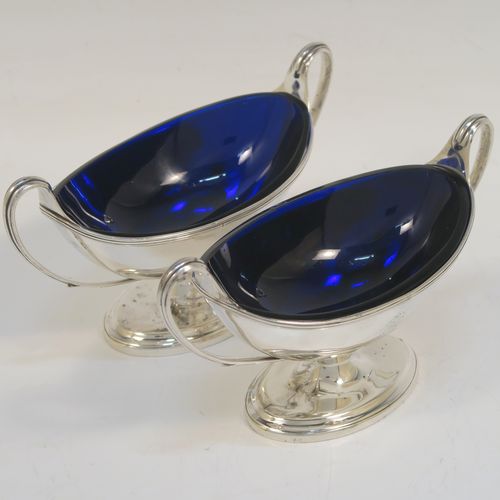 A very handsome Antique Georgian Sterling Silver pair of salt cellars, in a boat-shaped neoclassical style, having plain oval bodies, with blue-glass liners, applied reeded borders, and reeded side-handles, all sitting on pedestal feet. These elegant antique silver salt cellars were made by William Frisbee of London in 1792. The dimensions of these fine hand-made antique silver salt cellars are length 13.5 cms (5.3 inches), width 6 cms (2.3 inches), height 8 cms (3.25 inches), and they weigh a total of approx. 180g (5.8 troy ounces). Please note that both salt cellars have engraved crests.   