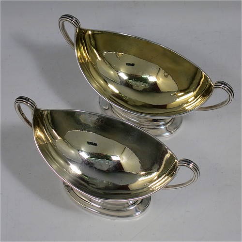 An Antique Georgian Sterling Silver pair of salt cellars, in a boat-shaped neoclassical style, having plain oval bodies, with gold-gilt interiors, applied reeded borders, and reeded side-handles, all sitting on pedestal feet. Made by John Wakelin and William Taylor of London in 1785 (having the rare Incuse duty mark). The dimensions of these fine hand-made antique silver salt cellars are length 13.5 cms (5.3 inches), width 6.5 cms (2.5 inches), height 8 cms (3.25 inches), and they weigh a total of approx. 214g (6.9 troy ounces). Please note that both salt cellars have engraved crests.